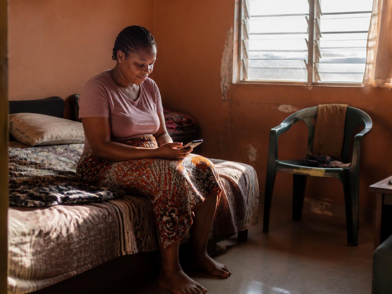 Uma mulher sentada na beirada da cama em um quarto em Goías, lendo no celular à luz do amanhecer